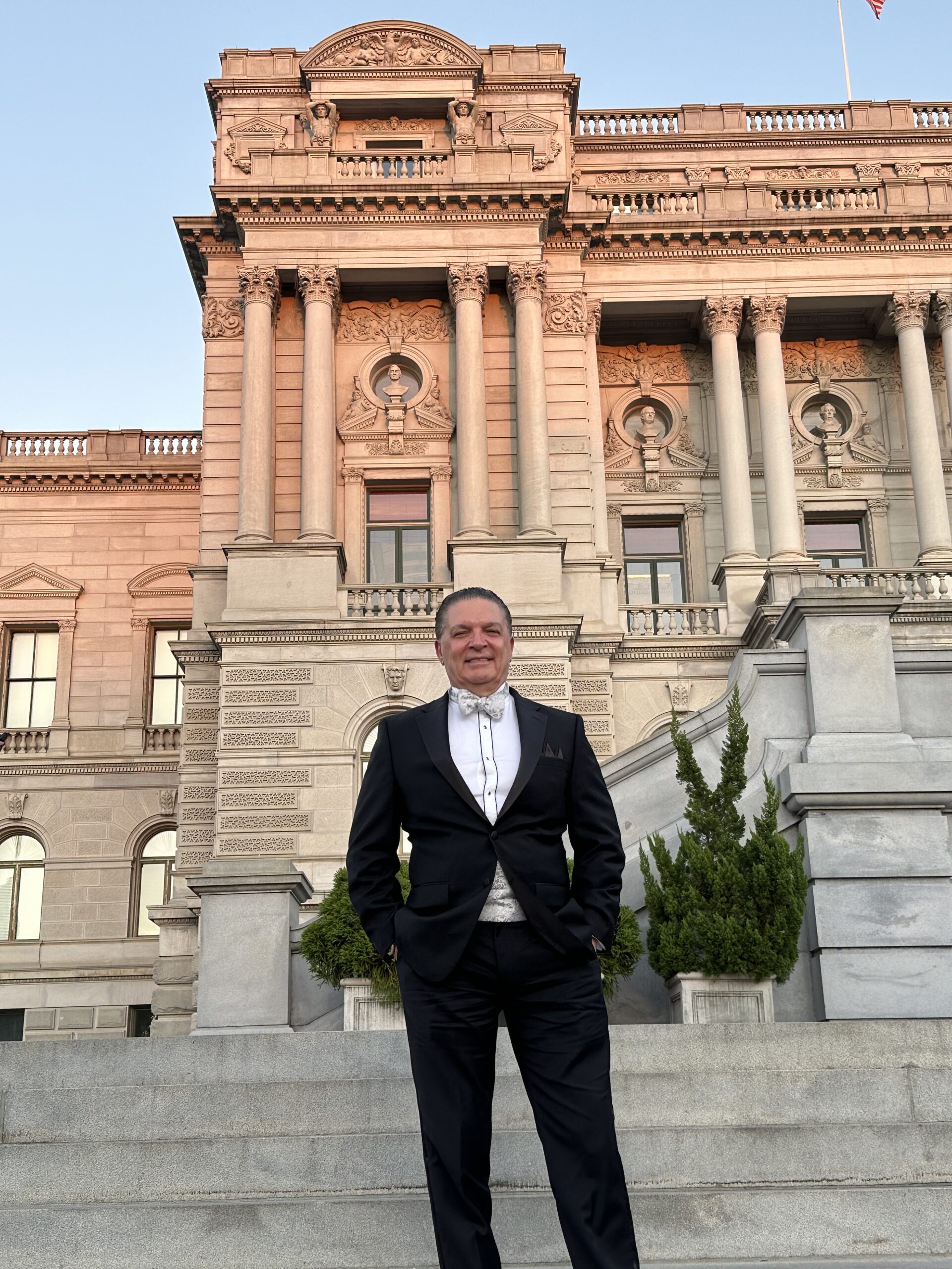 A man in a formal black suit and bowtie stands on stone steps in front of a building